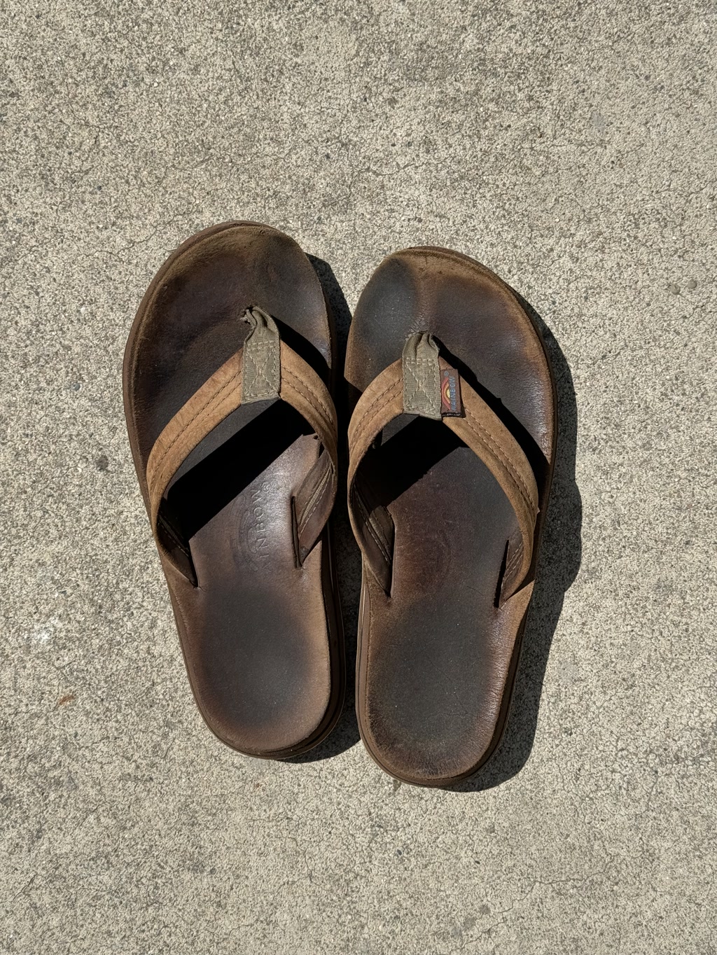 A pair of brown leather thong sandals are placed on a concrete surface. The sandals appear to have a slight wear and are designed with a single strap that splits into a Y shape forming the thong part that goes between the toes. There is a visible tag on the outer side of each sandal strap showing a small rectangular logo.