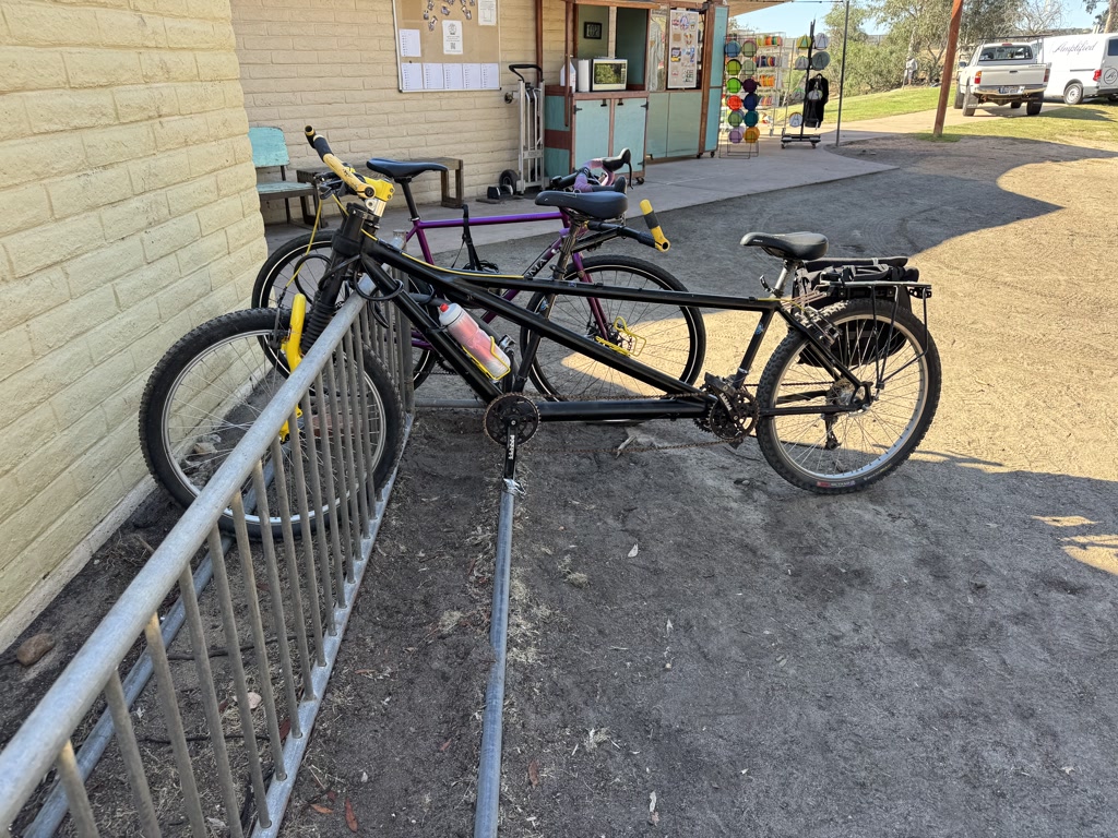 A tandem mountain bike is parked at a metal bike rack. The bike is primarily black with a yellow front fork. It has two seats and two sets of pedals, signifying it is built for two riders. Its features, such as the knobby tires and sturdy frame, suggest it is designed for off-road cycling. Further back, there is another bike leaning against the rack, pink and purple in color, with a water bottle attached. The setting appears to be a school or a public building, as indicated by children's artwork displayed on a board on the wall in the background. A vehicle is parked on the right, and the sky is clear, suggesting a bright day.