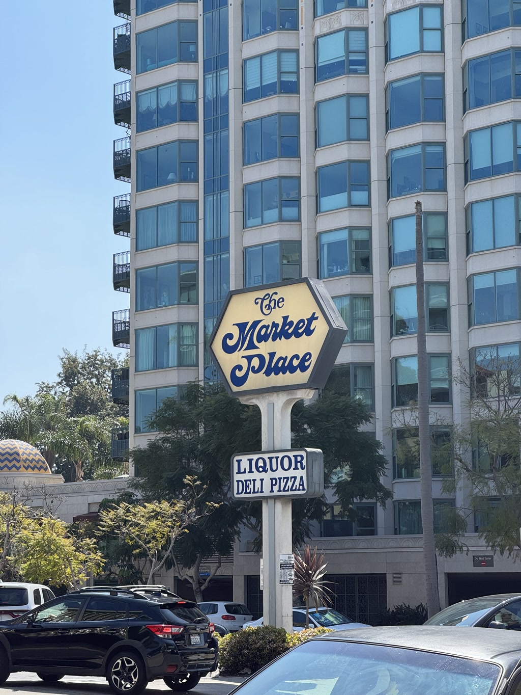A distinctive sign with an ornate, cursive font presents the name 'The Market Place' against a hexagonal backdrop with a dark blue border. Below it, a rectangular sign in a simpler, bold typeface lists the amenities available: 'LIQUOR DELI PIZZA.' The sign stands out in front of a multi-storied building with numerous balconies and reflective windows. Tall palm trees and a clear blue sky suggest a sunny, warm environment. Parked and moving cars indicate the presence of a busy street.