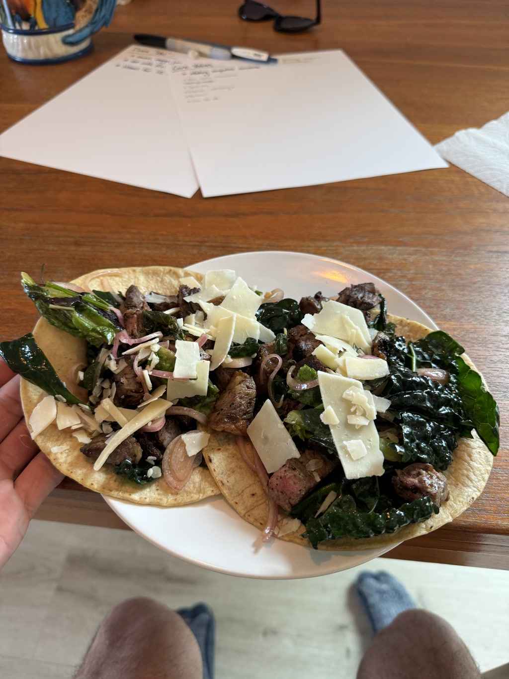 A plate is presented with what appears to be two open-faced tacos generously topped with slices of grilled steak, sautéed onions, greens, and shaved Parmesan cheese. The tacos rest on light-colored soft tortillas and are piled high with ingredients, suggesting a fresh and hearty meal. In the background, a wooden table surface is visible with two blank papers, a pen, and a pair of sunglasses laid out. No readable text is discernible.