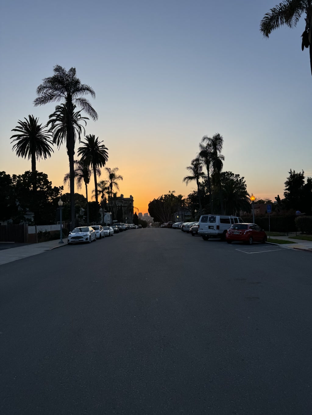 A serene dusky sky gradients from deep blue to a warm orange along the horizon. Tall palm trees line both sides of a street, standing out in silhouette against the twilight. The street is calm with parked cars on both sides, and the road stretches toward the setting sun. The environment is quiet, with no visible people or moving vehicles, suggesting a tranquil suburban setting in the evening.