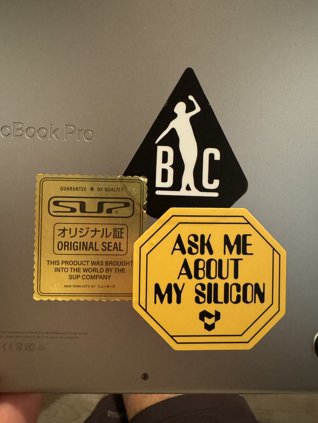 The bottom section of a MacBook Pro is shown adorned with three stickers. On the left is a golden rectangular sticker with stylized text and borders, resembling a vintage warranty seal. In the center is a triangular black sticker featuring a silhouette of a person with a hand reaching upwards, and the letters 'BC.' To the right is a yellow, hexagonal sticker with bold lettering that invites conversation about the owner's 'silicon,' alluding to the computer's processor. The laptop's aluminum surface has the model name embossed, and customary regulatory information printed at the bottom.