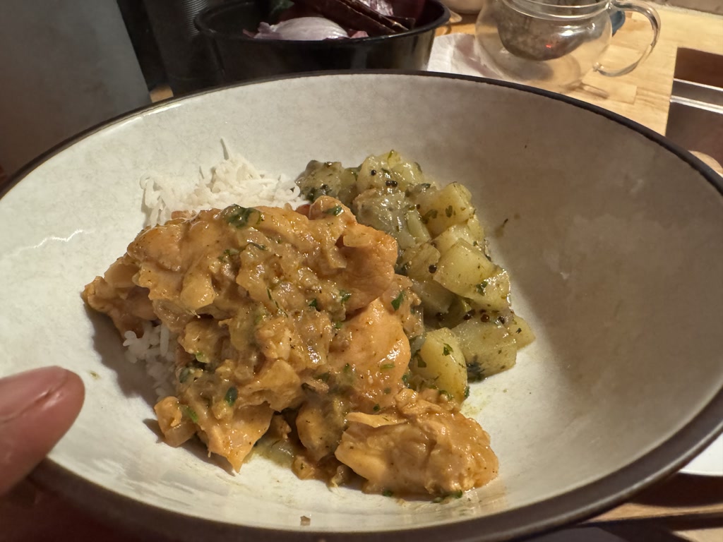 The photo displays a bowl containing two types of curry served on a bed of white rice. On one side of the bowl, there's a creamy, light orange-colored curry with chunks of what could be chicken, and it's garnished with small green flecks, possibly herbs. Beside it, there's a preparation of cubed vegetables that appear to be potatoes seasoned with green herbs. The bowl is perched in someone's hand, and the setting suggests an informal, homelike dining environment. There's a clear ambiance of a cozy meal.