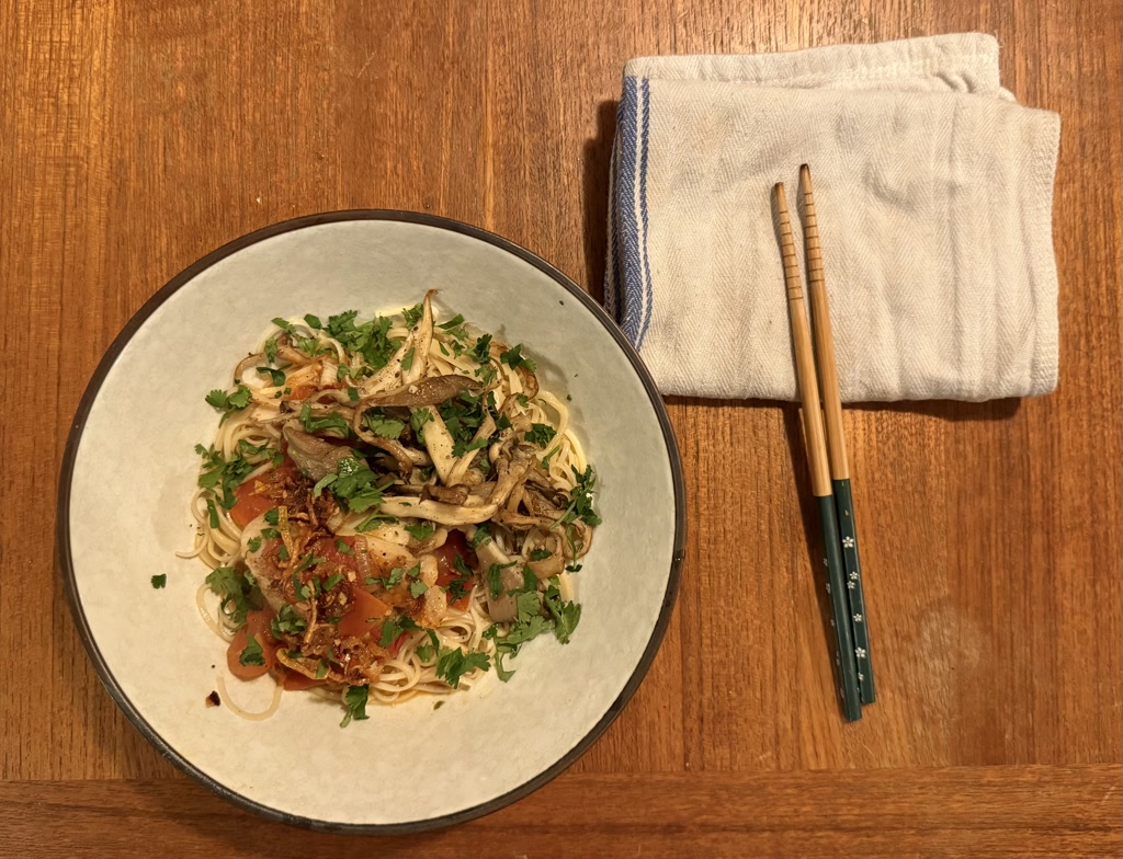 A bowl of homemade tomato poached cod with chili oil shallots on top, served over chuka soba noodles topped with garlic oyster mushrooms and garnished with lemon and cilantro. The dish is presented on a wooden table alongside a folded napkin and a pair of chopsticks with blue stars.