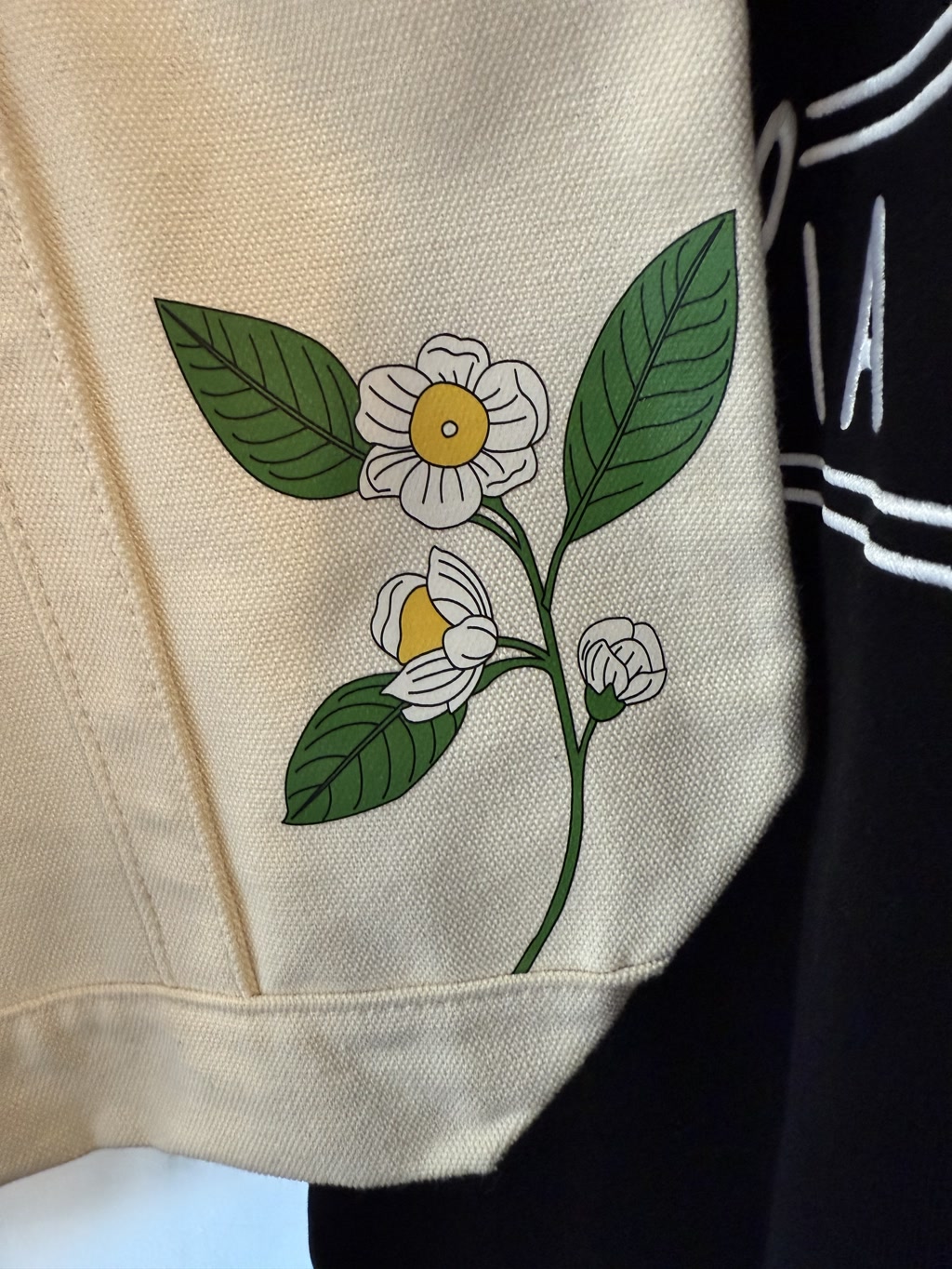 The image displays a close-up of a tote bag with a natural beige color. On the bag, there is a detailed illustration of a camellia plant. The drawing includes several green leaves and two white flowers, each with a yellow center. The design is elegant and simple, showcasing the beauty of the camellia. The tote appears to be made of a sturdy fabric, suitable for carrying various items.