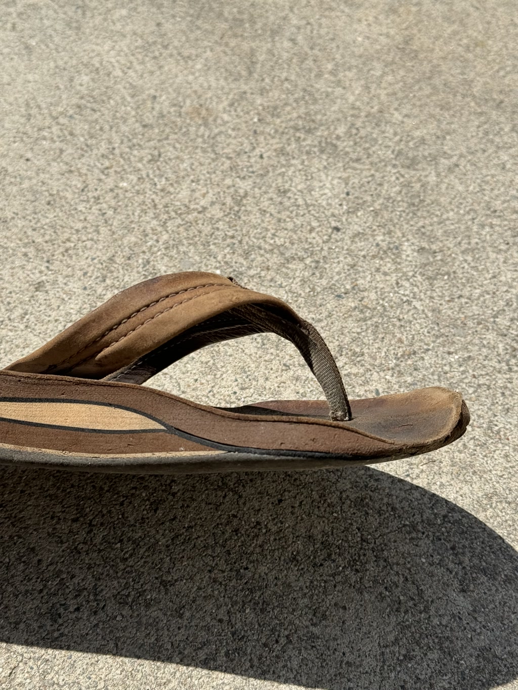 A single brown flip-flop with a dark brown strap is shown resting on a concrete surface. The flip-flop appears to be well-worn with signs of wear on both the sole and the footbed. The texture of the concrete is rough with small, scattered pebbles embedded in it. The flip-flop casts a sharp shadow on the ground, suggesting that the photo was taken in bright daylight.