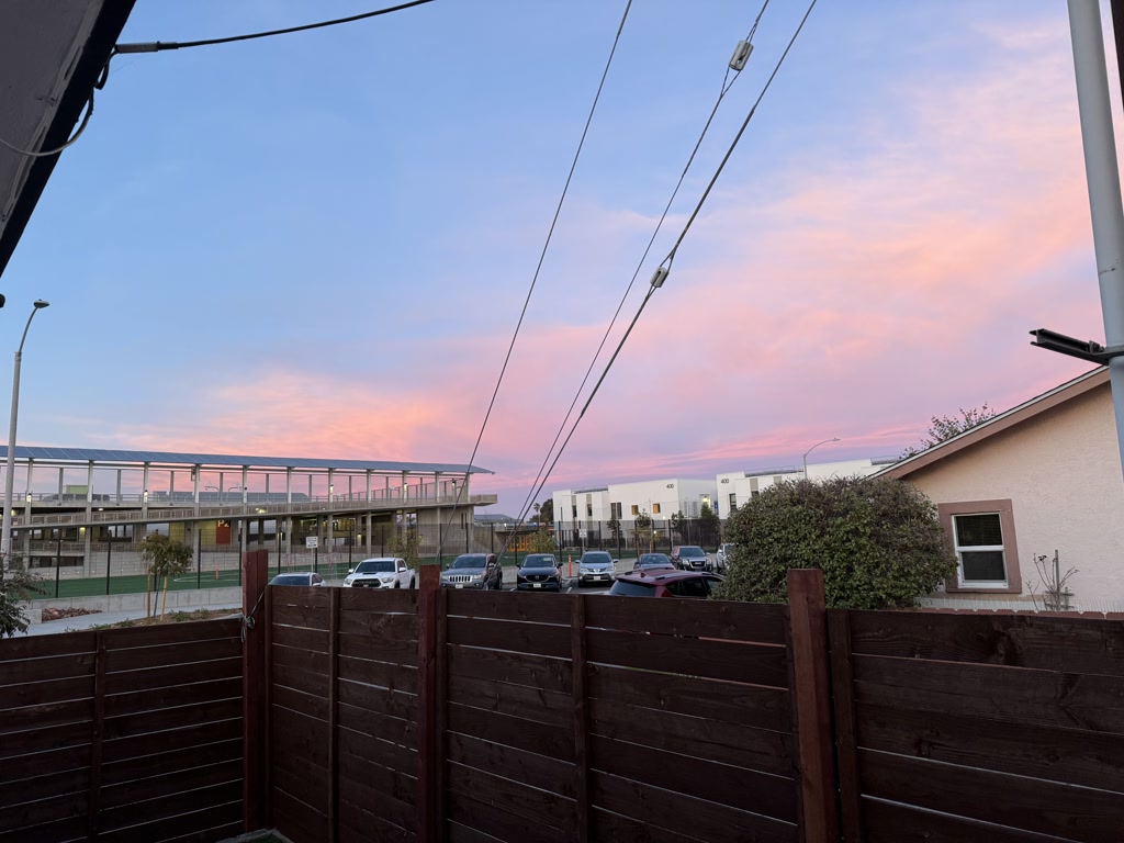 The image captures a tranquil evening scene with a stunning sunset painted in hues of pink and purple across the sky. The foreground features a wooden fence, behind which multiple vehicles are parked. To the left, there is a bridge with a glass railing, and a building with a modern design is visible in the background. Streetlights and other urban elements are also present, suggesting a suburban or urban setting. The vibrant colors of the sunset contrast beautifully with the muted tones of the buildings and vehicles.