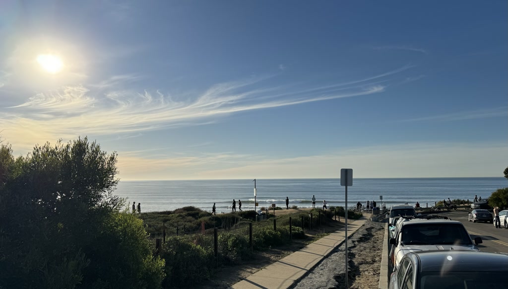 The scene captures a serene beach vista under a wide blue sky adorned with wispy cirrus clouds. The sun shines brightly in the upper left corner, casting a warm glow and a few gentle lens flares. A paved path on the right leads towards the beachfront, bordered by lush green shrubbery and a wooden fence, guiding visitors to enjoy the sands and surf. People are scattered across the frame, some by the water's edge and others milling around near parked cars lining the roadside. The horizon stretches across the middle, where the calm sea seamlessly meets the sky, creating a tranquil and inviting ambiance.