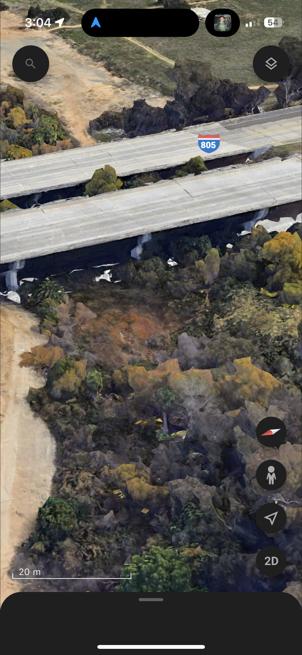 The view shows a three-dimensional, satellite perspective of a landscape where a freeway overpass, labeled '805', crosses above another road or area. The terrain looks rugged with patches of greenery and possibly some rocky formations. The image seems to have areas affected by digital rendering artifacts, giving some parts of the terrain a distorted appearance. User interface elements from a mapping application are visible, including search and location buttons, a scale indicator showing '20 m', and other navigation icons. The user interface also includes indications of the time '3:04', a navigation arrow, and some obscured elements at the top of the screen.