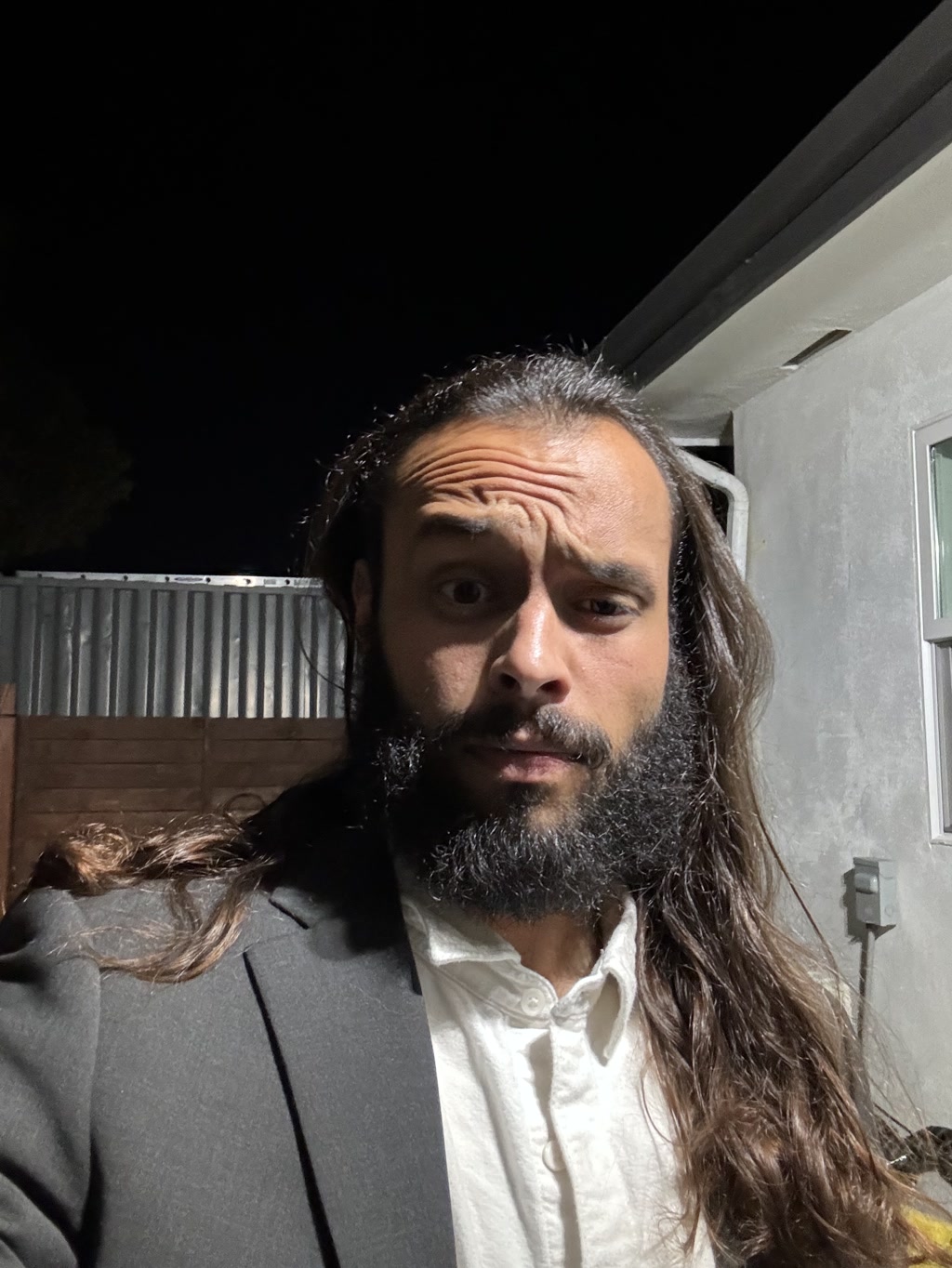 A person with long wavy hair and a full beard is capturing a selfie at night time. They are wearing a white collared shirt and a dark blazer. The background is dark with only the corner of a building and part of a wooden fence visible under a night sky.