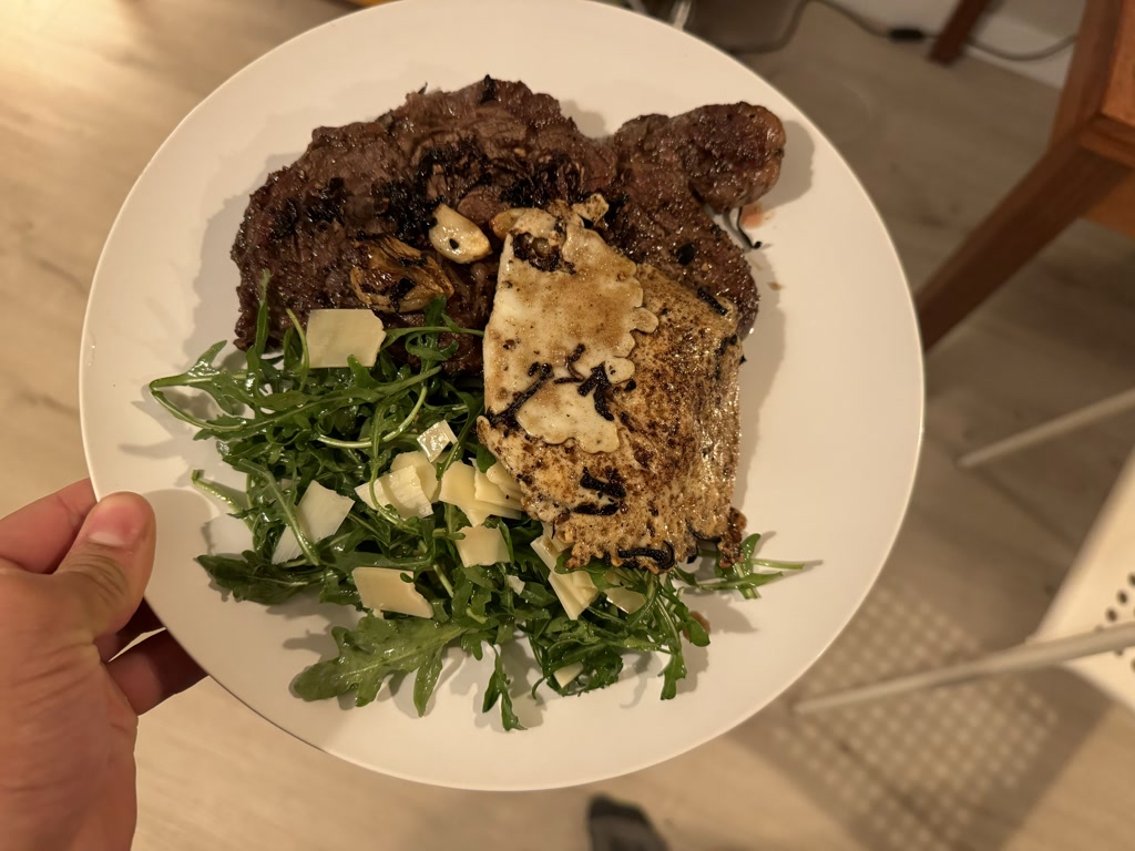 A plate holds a meal consisting of a seared steak, eggs over easy with a light brown, crispy edge, and a fresh arugula salad. The pieces of steak are cooked to a rich brown on the outside with visible grilling marks. The eggs appear tender with slightly runny yolks and are topped with sliced mushrooms. The salad is bright green, garnished with shaved parmesan cheese, offering a contrast in colors and textures to the other components of the dish.
