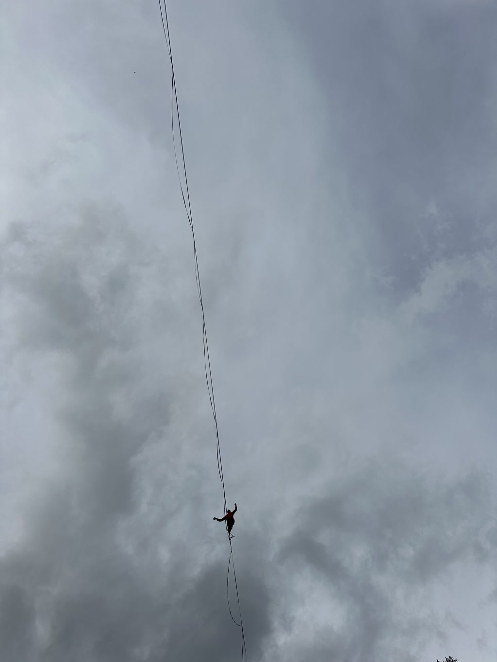 A person is balancing on a slackline that is stretched across a background of a cloudy sky. The slackline appears to be high above the ground and is not supported by any visible structures, suggesting that the person may be highlining, a form of slacklining performed at significant heights. The individual is standing with their arms outstretched to the sides, likely for balance. The sky is mostly overcast with gray and white clouds, signaling a gloomy or overcast weather.