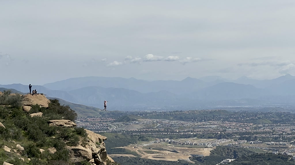 A person is walking on a slackline stretched between two rocky outcrops high above a sprawling cityscape. In the background, a mountain range looms under a cloudy sky. On the left side, a couple of individuals stand atop one of the outcrops, possibly securing or observing the slackliner. The vast urban area extends into the horizon with visible residential and commercial structures.