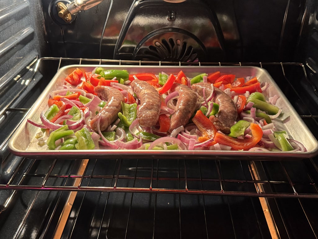 A tray of food in an oven, containing bourbon sausage, onions, and bell peppers, in the process of being roasted. The food is laid out on a flat surface, with sausages placed among slices of red and green bell peppers and chopped onions. The oven is on, and the tray is positioned inside it. The dish appears to be simple and hearty, suitable for a quick dinner.