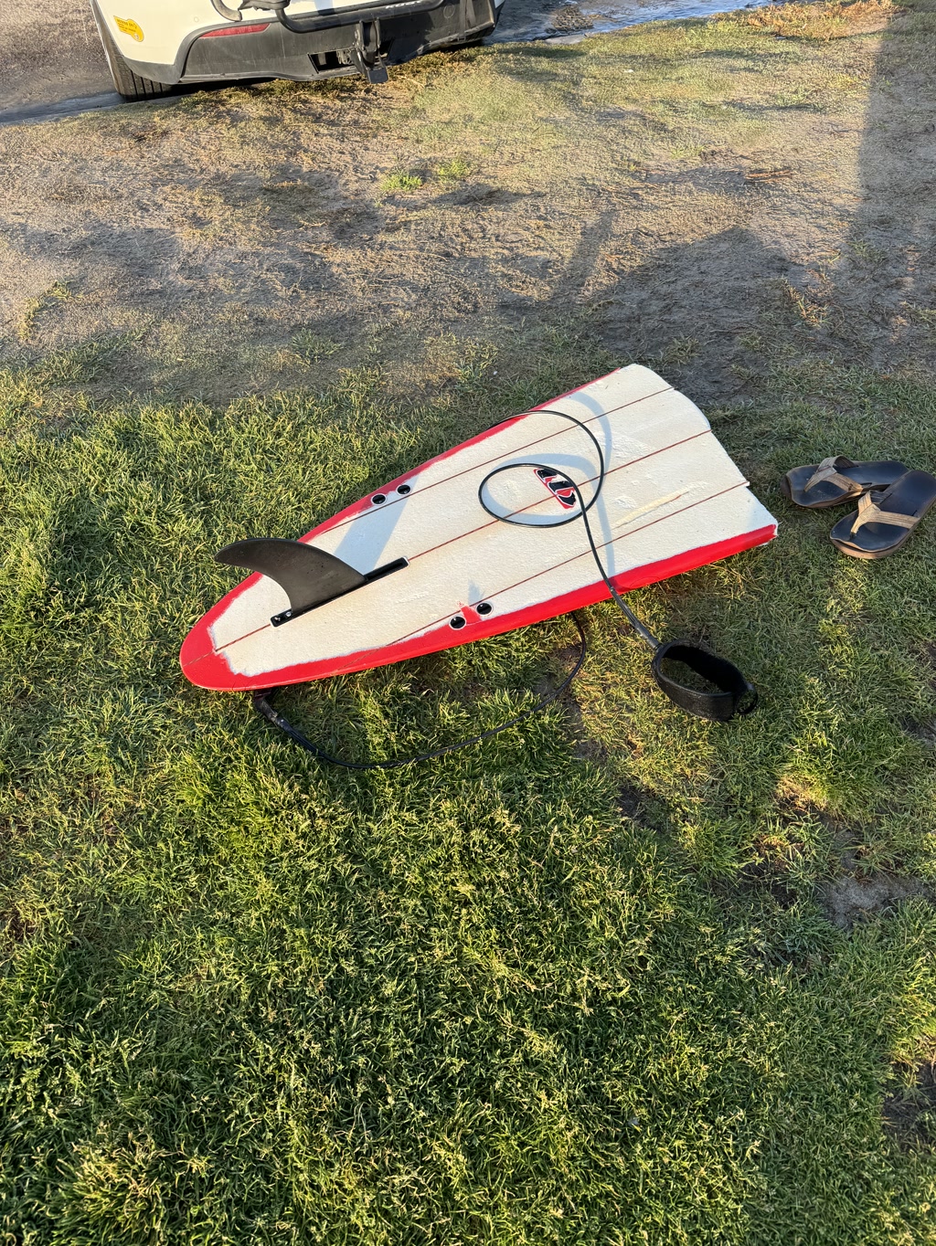 A red paddleboard with white borders is lying on the ground with the fin attached and ready for use. There is a black coiled leash attached to the board. Also visible are a pair of black flip-flops placed next to the board, and a black round object that appears to be a speaker or a container. Behind the board, there's a portion of a vehicle visible, which looks like the rear end of a van or SUV, showing the bumper, taillight, and a bit of the rear wheel. The ground shows a mix of grass and patches of dirt, suggesting this might be a transition area near a lake, river or beach used to enter or exit the water. The setting is illuminated by bright sunlight casting shadows on the ground, indicating it's either morning or late afternoon.