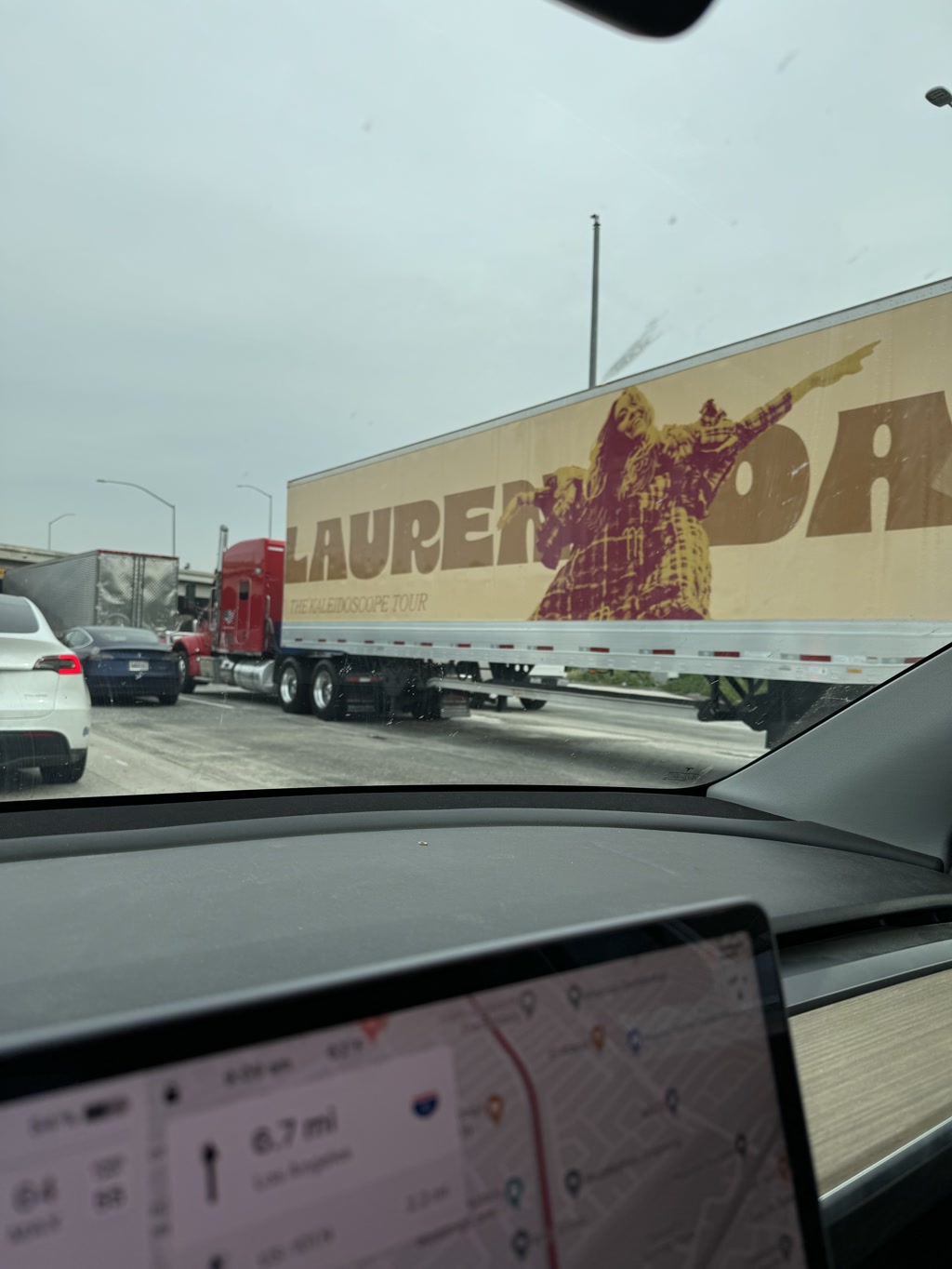 A red semi-truck is pulling a large trailer with a graphic and text printed on its side, featuring a human figure in a dynamic pose with one arm stretched out. The background has a warm tone, suggesting a psychedelic design. Behind the truck, there are other vehicles on the road, indicating a highway or busy street setting. The view is from inside a vehicle with part of the dashboard visible, including a navigation screen that displays a map and some driving metrics.