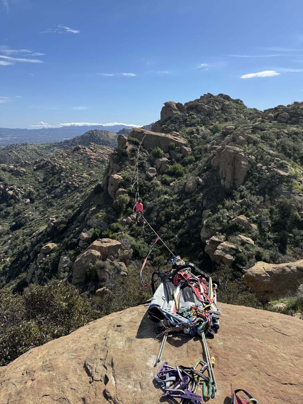 A collection of rock climbing equipment is arranged on a flat rock surface with a stunning mountainous landscape in the background. The equipment includes carabiners, slings, and other climbing hardware, suggesting preparation for a climb or the conclusion of one. A climbing rope extends from the gear, reaching towards another rocky outcrop in the distance. The landscape features rolling hills with sparse vegetation under a clear blue sky dotted with a few wispy clouds.