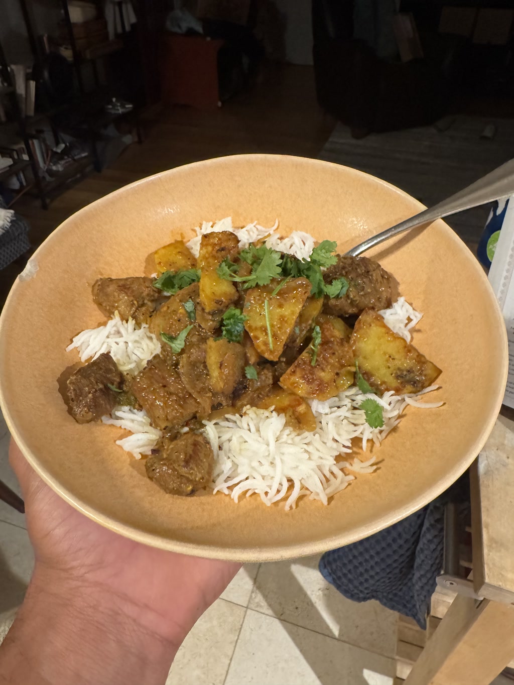 A plate of food consisting of rice topped with chunks of meat and pieces of golden-brown fried potatoes, garnished with fresh green coriander leaves. The dish appears to be a curry, likely a vindaloo, which is a Portuguese-Indian cuisine item made with meat traditionally marinated in vinegar and garlic. The plate being held shows a generous portion of the meal that looks appetizing with its savory and spicy components combined.