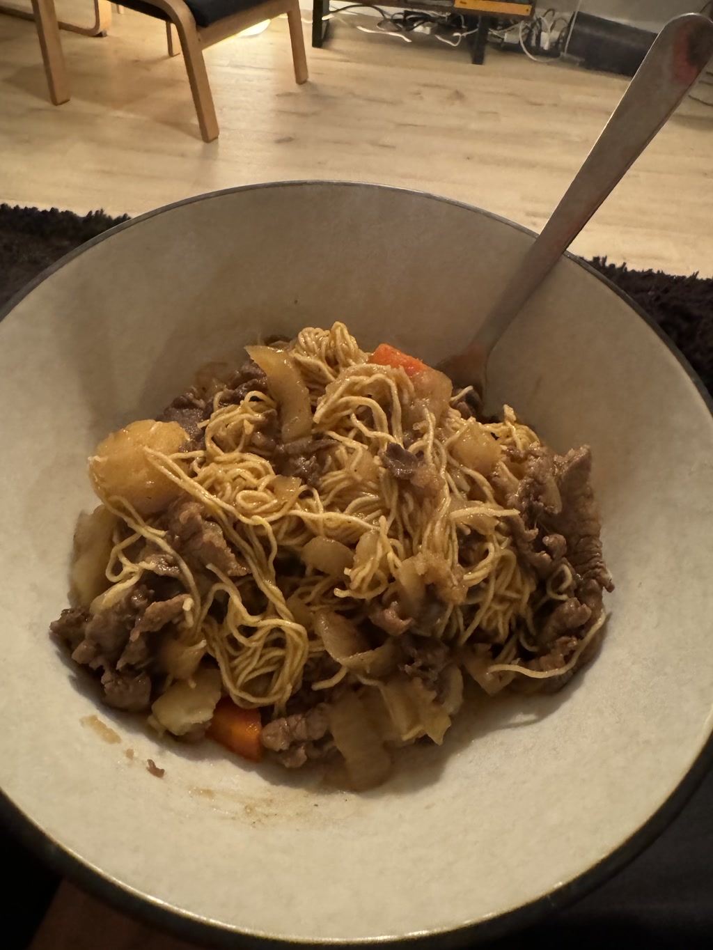 A bowl filled with a stir-fried noodle dish is present, featuring what appears to be thin noodles, chunks of beef, and possibly some vegetables like onions and carrots. There is a distinct shine to the ingredients suggesting they may be coated in a sauce or oil. The bowl is sat on a surface with a fork resting inside, indicating the meal is ready to be eaten. The backdrop is a casually set room with chairs and a rug, partially showcasing an indoor domestic setting.