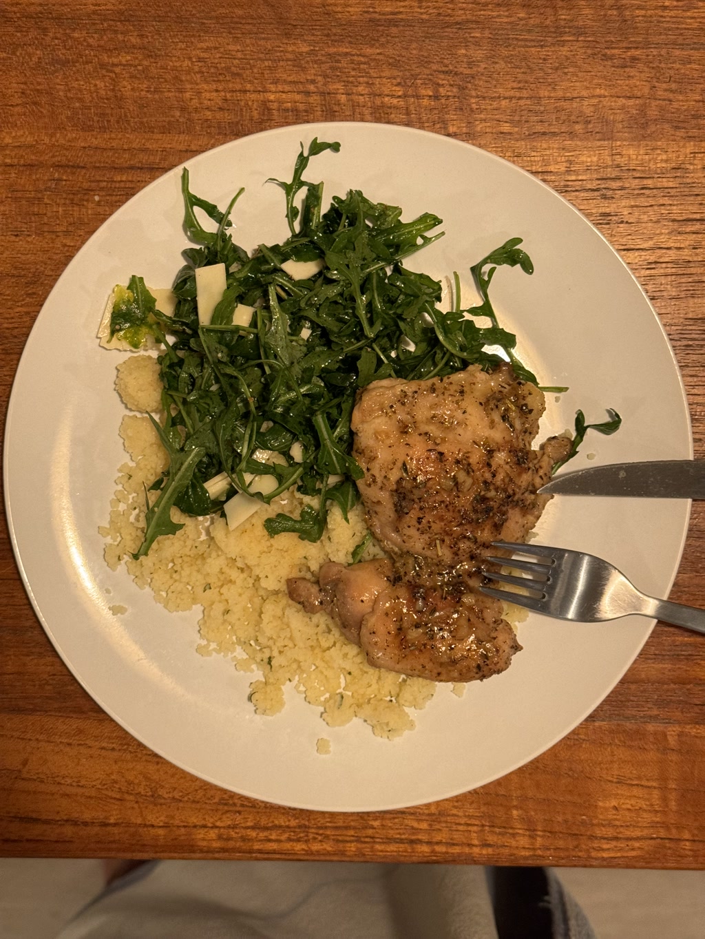 A plate of food featuring fire garlic miso chicken, a light brown piece of chicken seasoned with visible spices, accompanied by a fluffy bed of pale yellow couscous, and a side of arugula salad dressed and mixed with what appears to be pieces of parmesan cheese, adding hints of white to the green leaves. The food is presented on a round white plate, resting on a wooden table. A fork is placed to the right of the chicken, partially resting on the couscous. The plate offers a wholesome and nutritious meal with a balance of protein, grains, and greens.