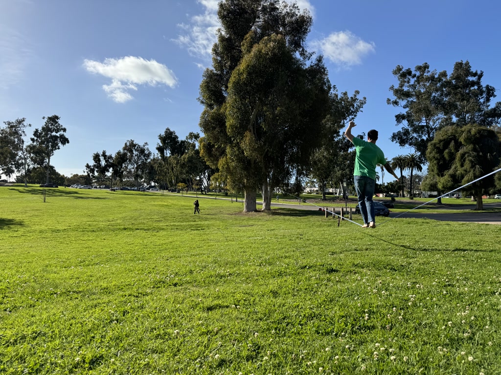 Ryan is enjoying his time balancing on a slackline stretched between two points in a vibrant, grassy park. The scene depicts a clear sky with a few scattered clouds and a rich variety of trees in the background, adding to the serene atmosphere. There's a small figure in the distance, which appears to be another person in the park, potentially spectating or engaged in their activity. Cars are visible parked along the road, which borders the park area.