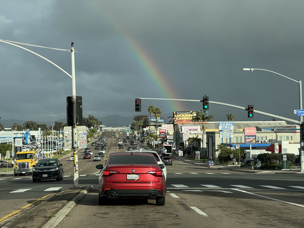 A view from a car stopped at a traffic light, looking down a bustling urban street with vehicles in both directions. The sky appears stormy with dark clouds, but there's also a visible rainbow on the left side of the street. Numerous signs from businesses and billboards line the roadside, advertising a casino, car rental companies, and automotive services among others. On the right-hand side, an Avis and Budget car rental sign can be seen. Traffic lights, street lamps, and power lines cross over the street.