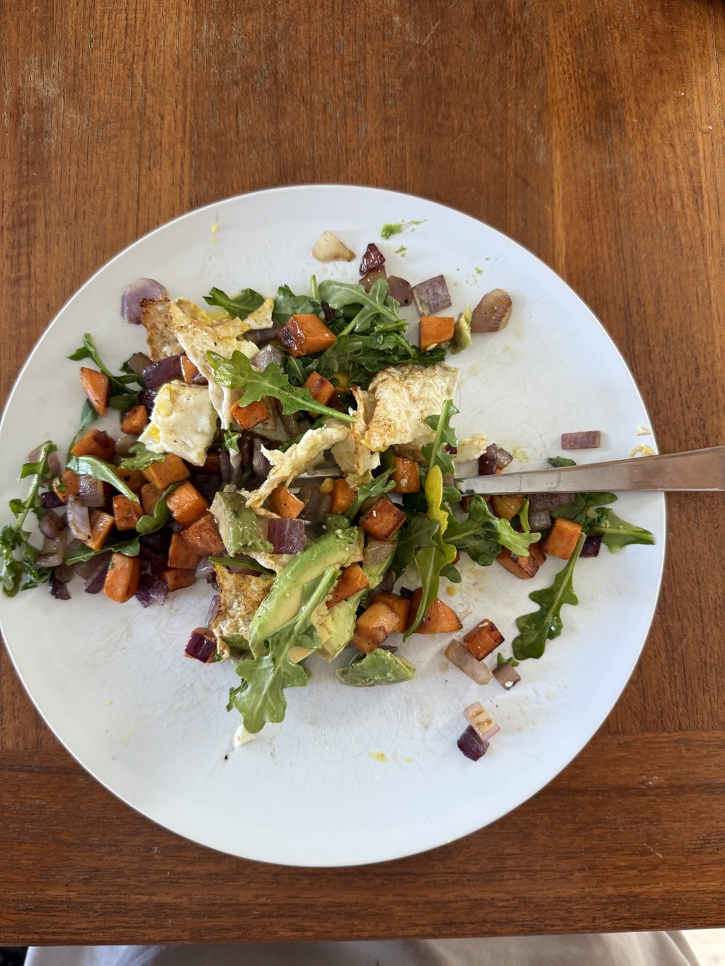 A delicious breakfast plate featuring a small sweet potato hash with diced sweet potatoes, arugula, eggs, avocado, and onion. Served on a white plate with a fork on the side, placed on a wooden table.