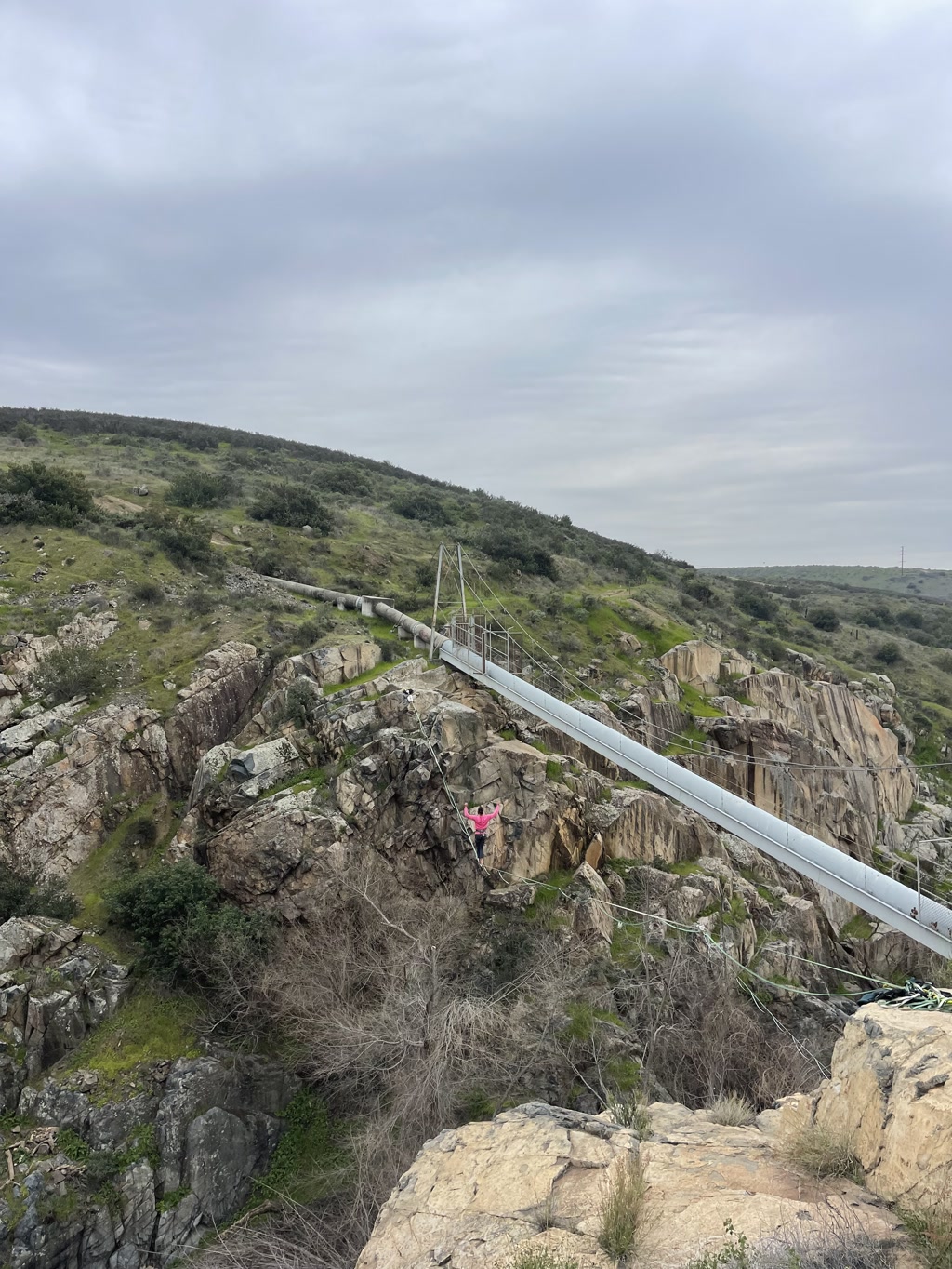 A suspension bridge spans across a rugged terrain of steep, weathered rocks and sparse greenery. Below the bridge, a person wearing bright pink is seen mid-climb on the rocky cliff face, adding a pop of color to the natural landscape. Overcast skies loom above, and no text is visible in the scene.