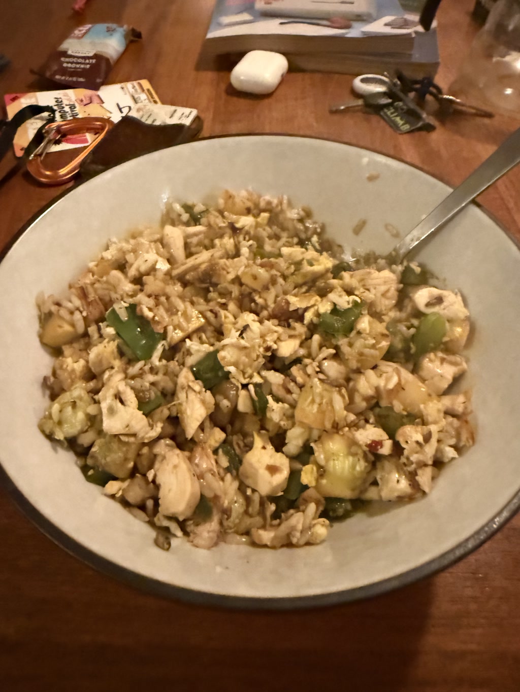 A bowl of simple chicken fried rice with bell peppers and squash. The dish is served in a light-colored bowl with a silver spoon inserted into it. In the background, there are various items including a book, a marshmallow, a pair of keys, and some packets, suggesting a casual dining setting.