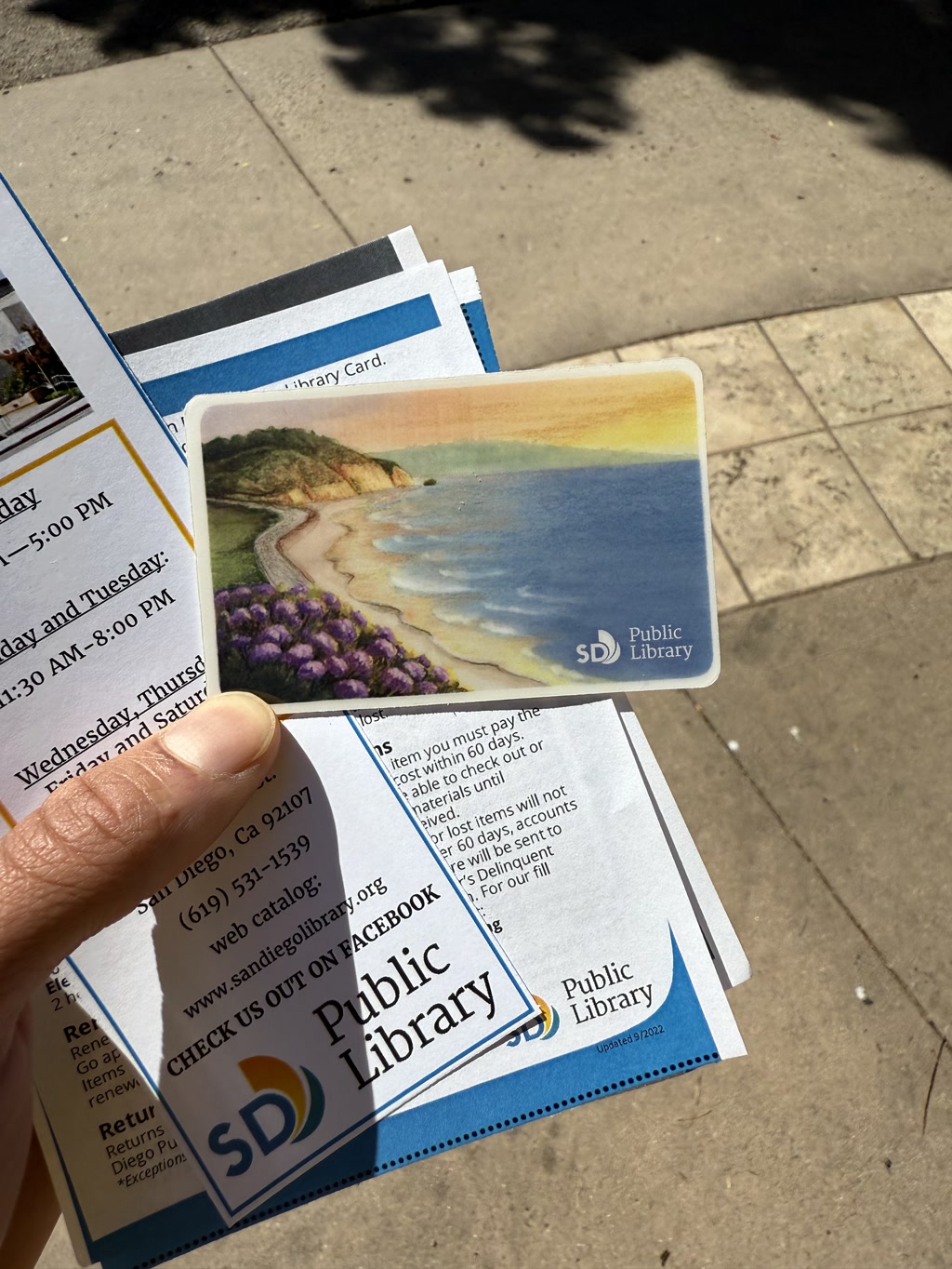 A hand holds a bundle of materials related to a public library. The items include a card with an artistic illustration of a coastal landscape featuring a sandy beach, purple flowers, hills, and a vibrant sunset. The stack also contains several printed papers, including one with details about library hours and another about returning items. The materials are branded with 'SD Public Library' and include a web catalog address and a Facebook page invitation. The context suggests an outdoor setting with sunlight and tree shadows on the ground.