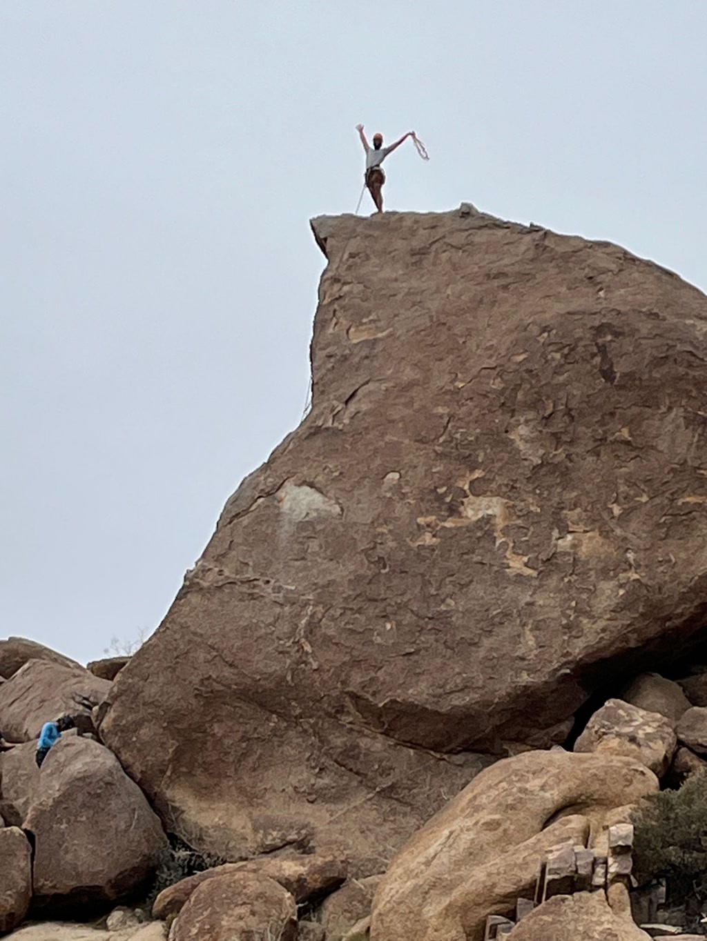 A person stands triumphantly on top of a large, smooth rock formation that resembles an elf's hat, set against a hazy sky. They appear to be celebrating their climb with a victory pose, arms raised high holding a loop of rope. Another individual is seen near the bottom left of the rock, dressed in blue, looking up towards the person at the summit.