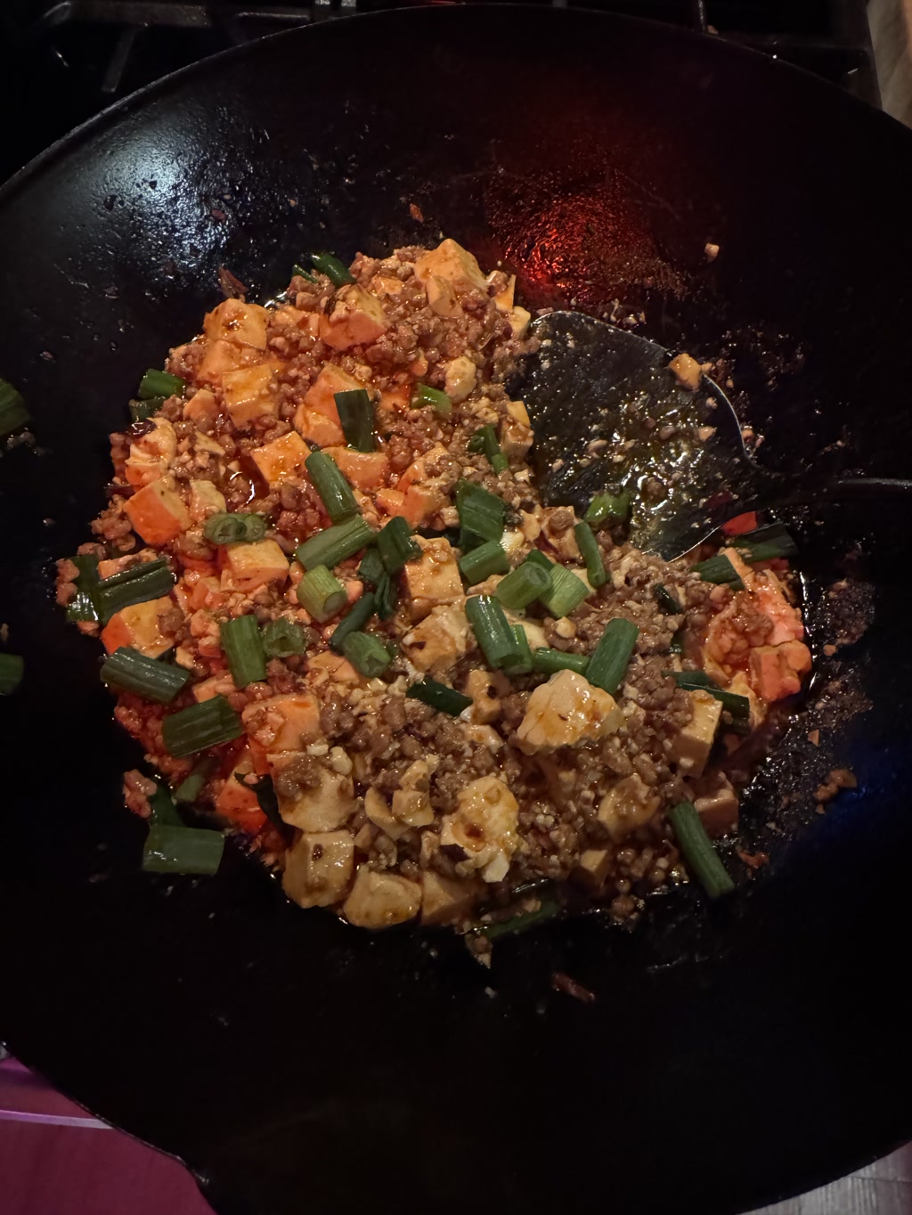 The image shows a close-up of a wok containing a dish that appears to be Mapo Tofu. The dish consists of chunks of tofu mixed with ground meat, typically pork or beef, and garnished with chopped green onions. The tofu chunks are coated in a spicy, dark red sauce, indicative of the traditional Sichuan flavors. The wok is black and has a metallic spoon in it, suggesting the dish is freshly cooked. The ingredients are evenly distributed, and the vibrant colors of the tofu, meat, and green onions create a visually appealing mix.