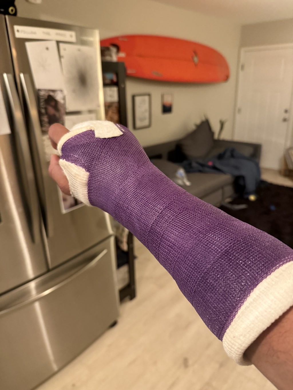 A purple armored gauntlet with a metal plate on the back of the hand and a finger hole is shown against the background of a modern kitchen with a silver refrigerator and various magnets and notes on it/