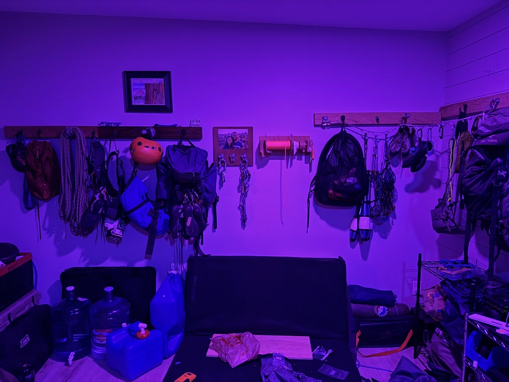 A room featuring a variety of climbing gear and equipment neatly organized on a wall. The gear includes harnesses, helmets, carabiners, ropes, and bags among other items. A climbing helmet is prominently displayed alongside the equipment. The wall features wooden panels and a couple of picture frames. Below the gear, there are some water jugs, a container of disinfecting wipes, and more equipment strewn across the floor, including a folded chair and various bags. The room is illuminated with purple lighting which casts a vivid hue over everything.