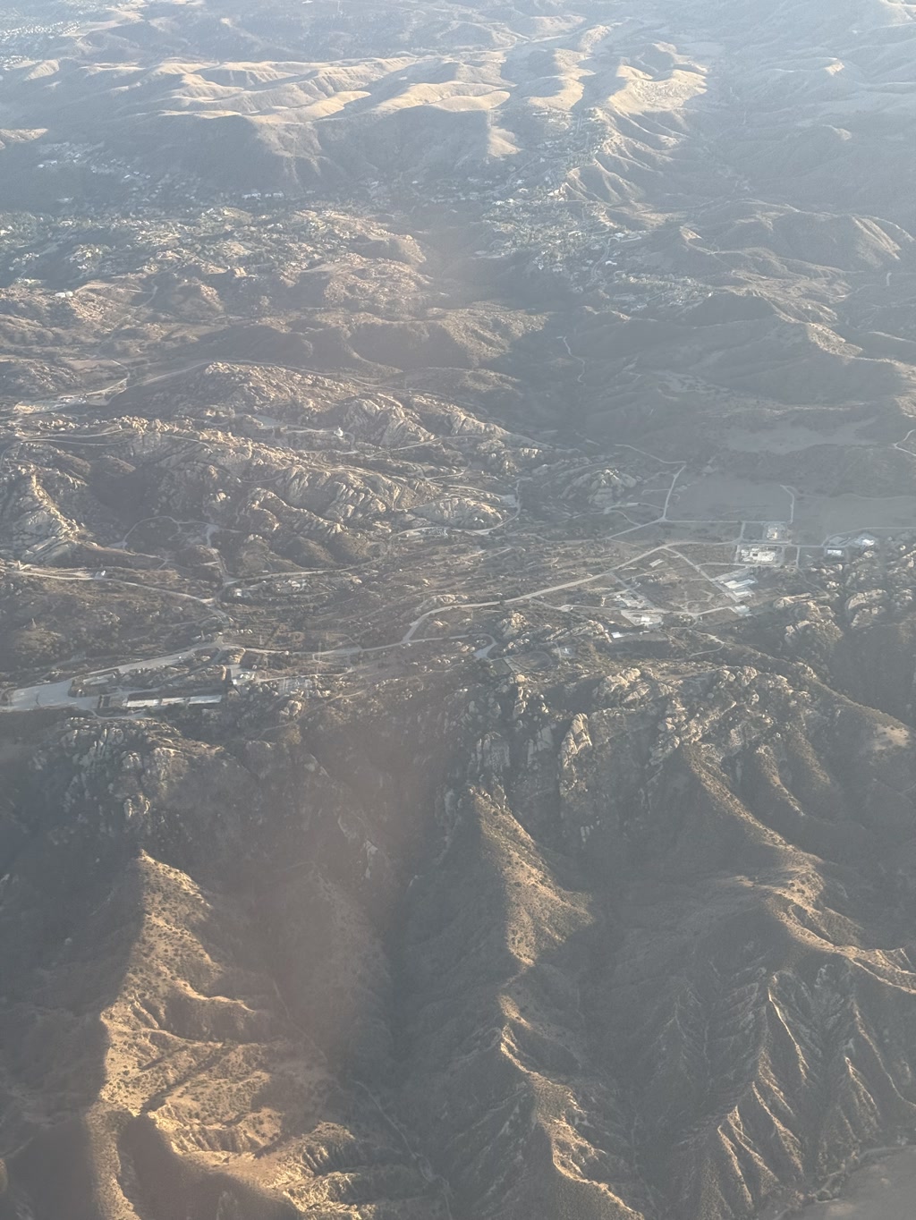 A stunning aerial view of a mountainous region, showcasing rugged, sunlit peaks and vast valleys. The landscape features winding roads and scattered settlements, giving a sense of isolation and natural beauty. The contrast between the shadows and the illuminated slopes adds depth and drama to the scene.