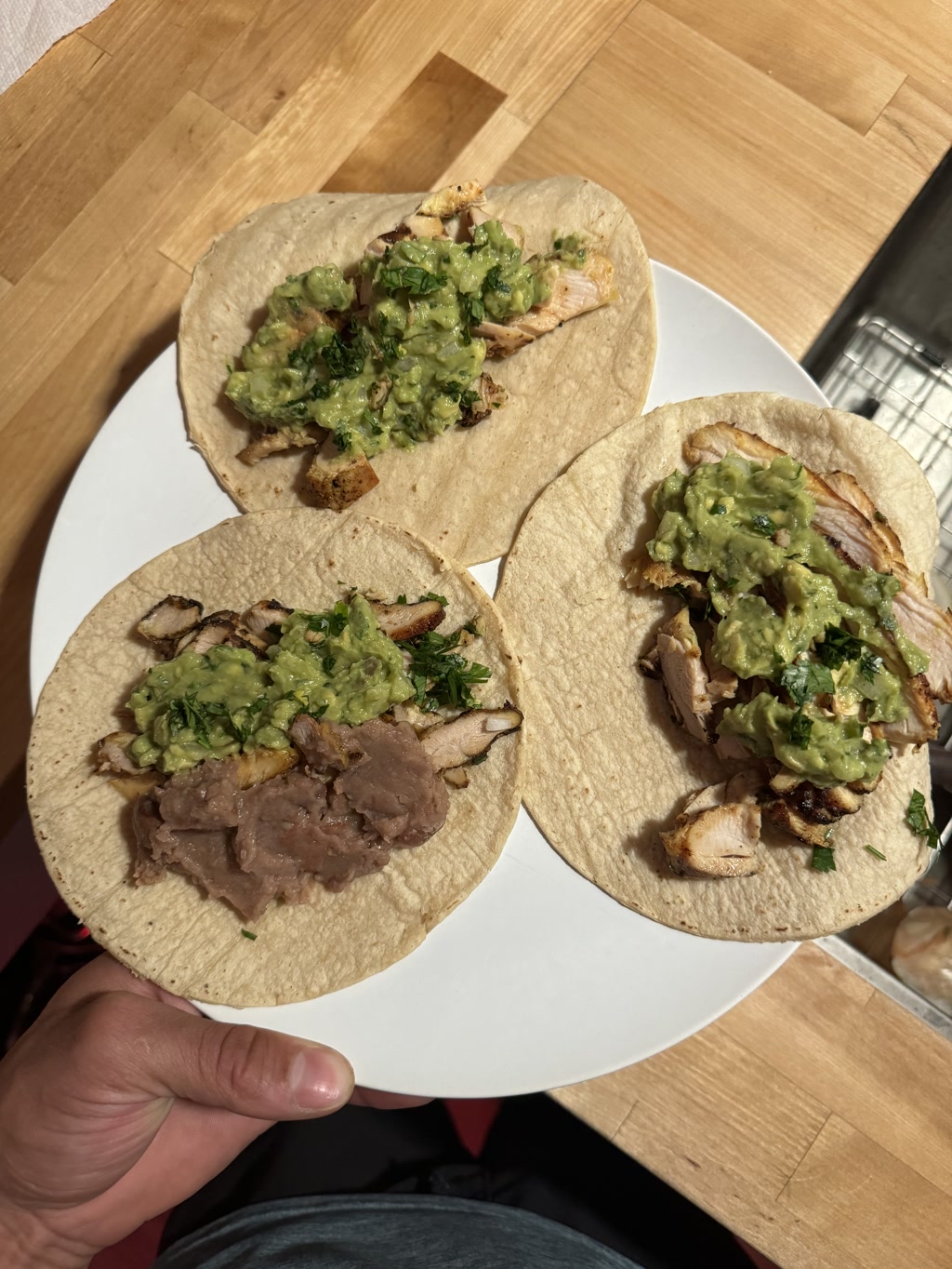 Three corn tortillas are topped with various ingredients. The first one has a layer of refried beans adjacent to some chopped herbs. The second and third tortillas are filled with grilled chicken pieces topped with a generous portion of chunky green salsa or guacamole. All are positioned on a white plate, which is being held by a person with visible fingers and a thumb. The plate rests on a wooden surface partially lit by sunlight, indicating either outdoor dining or the presence of a window nearby.