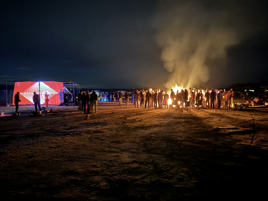 A night time gathering of people around a bonfire, with a vibrant blue and red illuminated geometric canopy in the background. The scene is set on a flat expanse with a distinctly barren ground, suggesting an outdoor festival or camping event. The canopy appears to be part of a temporary structure, likely a stage or art installation, which is glowing under the night sky. The fire is blazing with high flames and emitting a significant amount of smoke that rises into the dark sky. Various individuals are visible, some standing and talking, others sitting on the ground, contributing to a casual and social atmosphere. No close-up details of individuals can be discerned, preserving their anonymity.