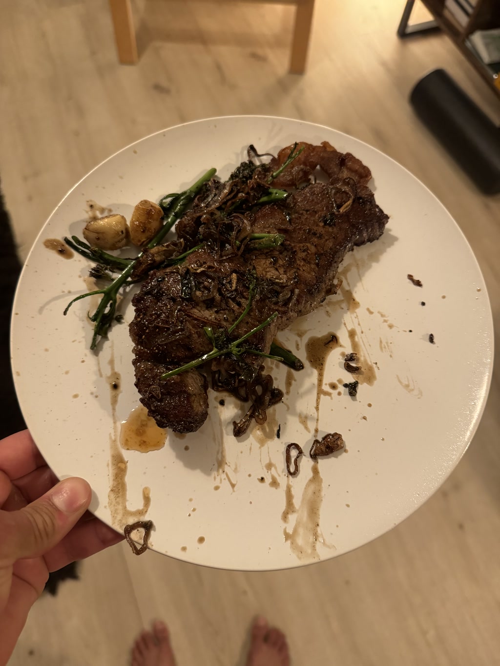 The photo shows a cooked steak with browned edges and visible grilling marks, suggesting it might be pan-seared or grilled. Accompanying the steak are some roasted garlic cloves and sprigs of a green herb, possibly rosemary, which are scattered on and around the meat. Also present are some fried crispy elements that could be onion or shallots. There are a few splashes and droplets, likely oil or a sauce, on the plate, indicating the steak may have been recently cooked or dressed. In the background, partially visible is a person holding the plate, with bare feet standing on a wooden floor, indicating an informal and personal setting. There's a hint of furniture in the far background, suggesting the photo was taken indoors in a living or dining area.