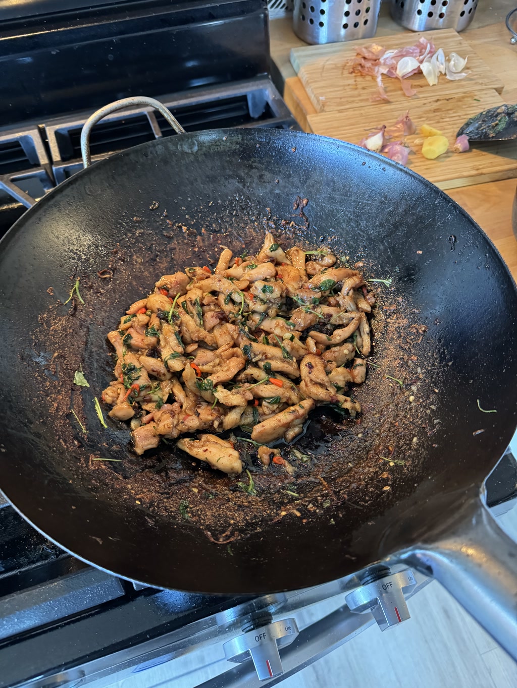 Stir-fried ingredients are in a wok on a gas stove, with visible heat controls at the bottom. The wok contains what appears to be chicken pieces cooked with a variety of herbs and spices, some of which have blackened from the high heat of the wok. The herbs may include green leaves like cilantro or parsley. There is evidence of browning on the sides of the wok, suggesting thorough cooking or sautéing. In the background, there is a wooden cutting board with remnants of chopped red onion and garlic skins indicating prior preparation.
