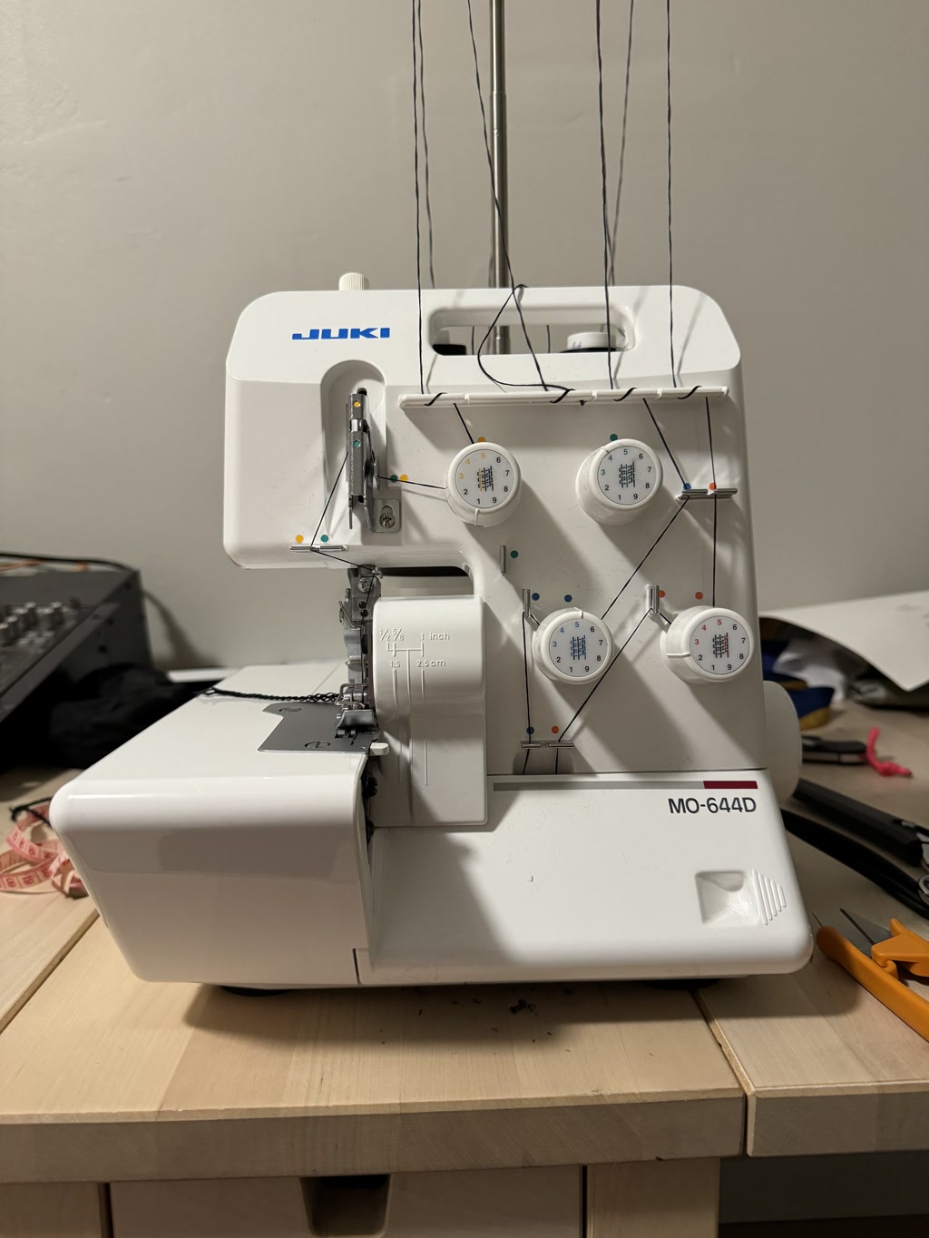 A Juki MO-644D model overlock sewing machine is pictured displaying four spools of thread mounted on top, with threaded paths leading down to the machine. The machine is made of white plastic and has three dials on the front, each with different settings for stitch functions, and measurement guides next to the needle plate indicating quarter inch and 1.5 cm seam allowances. There are scissors and a measuring tape scattered in the background indicating a sewing workspace.