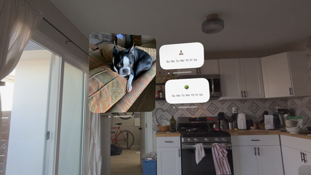A comfortable, well-lit room featuring a kitchen with white cabinetry and a patterned backsplash. On the countertop, various kitchen appliances, including a coffee maker, are visible. A bicycle is parked in the room, indicating a lifestyle that includes physical activity. The space is overlaid with augmented reality (AR) bubbles – one showing a Boston Terrier dog resting on a couch, symbolizing time spent with friends' pets, and two other bubbles each displaying a calendar icon and a meditation icon, indicating scheduled times for outdoor activities and meditation sessions.