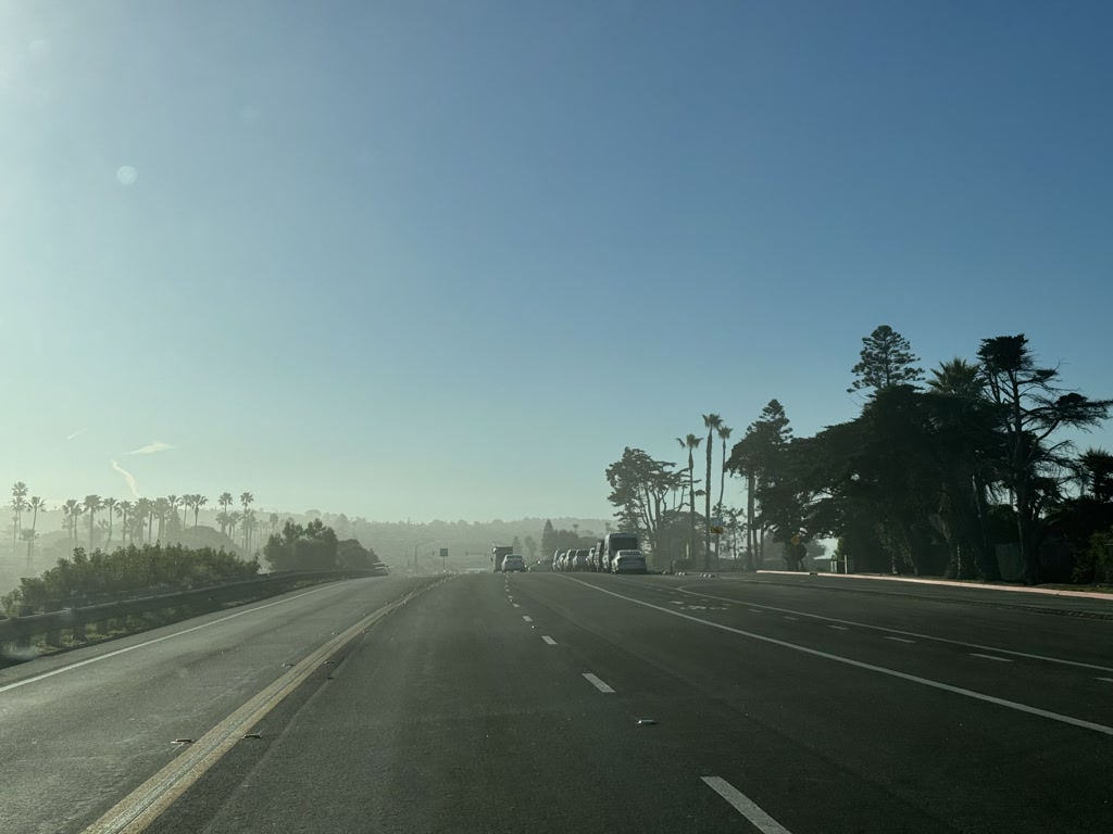 The scene captures a wide road with multiple lanes under a clear blue sky, with just a few wispy clouds. The road is fairly empty with only a few vehicles in sight, spread out across the lanes. On the right, there's a line of tall palm trees punctuating the landscape, suggesting a location with a warm climate. The horizon in the distance is hazy, which could indicate some fog, mist or just a brightness in the sky due to the angle of the sun. Roadside infrastructure such as lamp posts and traffic signs are also visible.