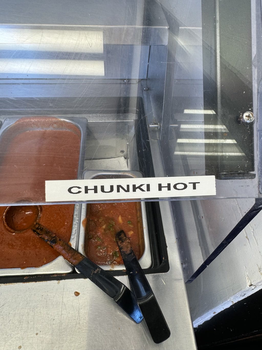 The photo shows two containers within a metal serving station, presumably at a food establishment. The container on the left appears to contain a smooth, mildly textured red sauce. The container on the right holds a chunkier sauce with visible green herbs and red base, likely some type of salsa. Both containers are equipped with black serving ladles that have stems exhibiting signs of frequent use indicated by their discolored and worn look. There is a label fixed above the containers with a text indicating the nature or name of the chunkier sauce.
