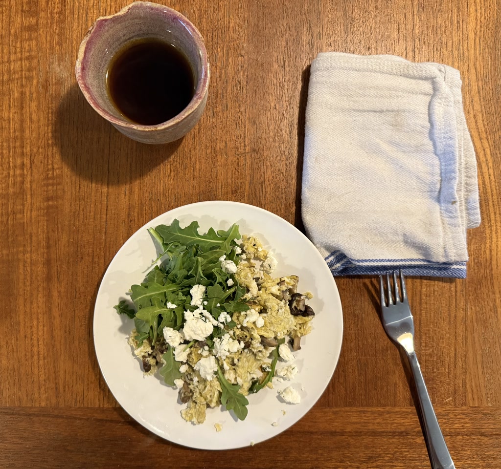 The image shows a breakfast setup on a wooden table. There is a plate with scrambled eggs mixed with arugula and feta cheese. A fork is placed next to the plate, and there is also a blue and white napkin. Additionally, there is a ceramic cup filled with dark liquid, likely coffee or tea.