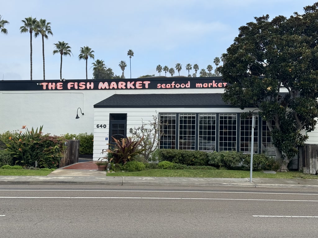 A white single-story building with a black roof is situated in front of a clear sky dotted with clouds. A line of tall palm trees stands in the background, creating a tropical scene. The establishment is named 'The Fish Market', as indicated by prominent red and black lettering on its facade. Segmenting the name, 'THE FISH' is on one side of a graphic outline of a fish, with 'seafood market' on the other. The entrance features a white door framed by large windows on both sides, and is accessible via a concrete pathway flanked by lush greenery and flowering plants. The building number, 640, is displayed next to the door. A street runs parallel to the front of the property, marked with a solitary street sign and a clear pedestrian crossing.