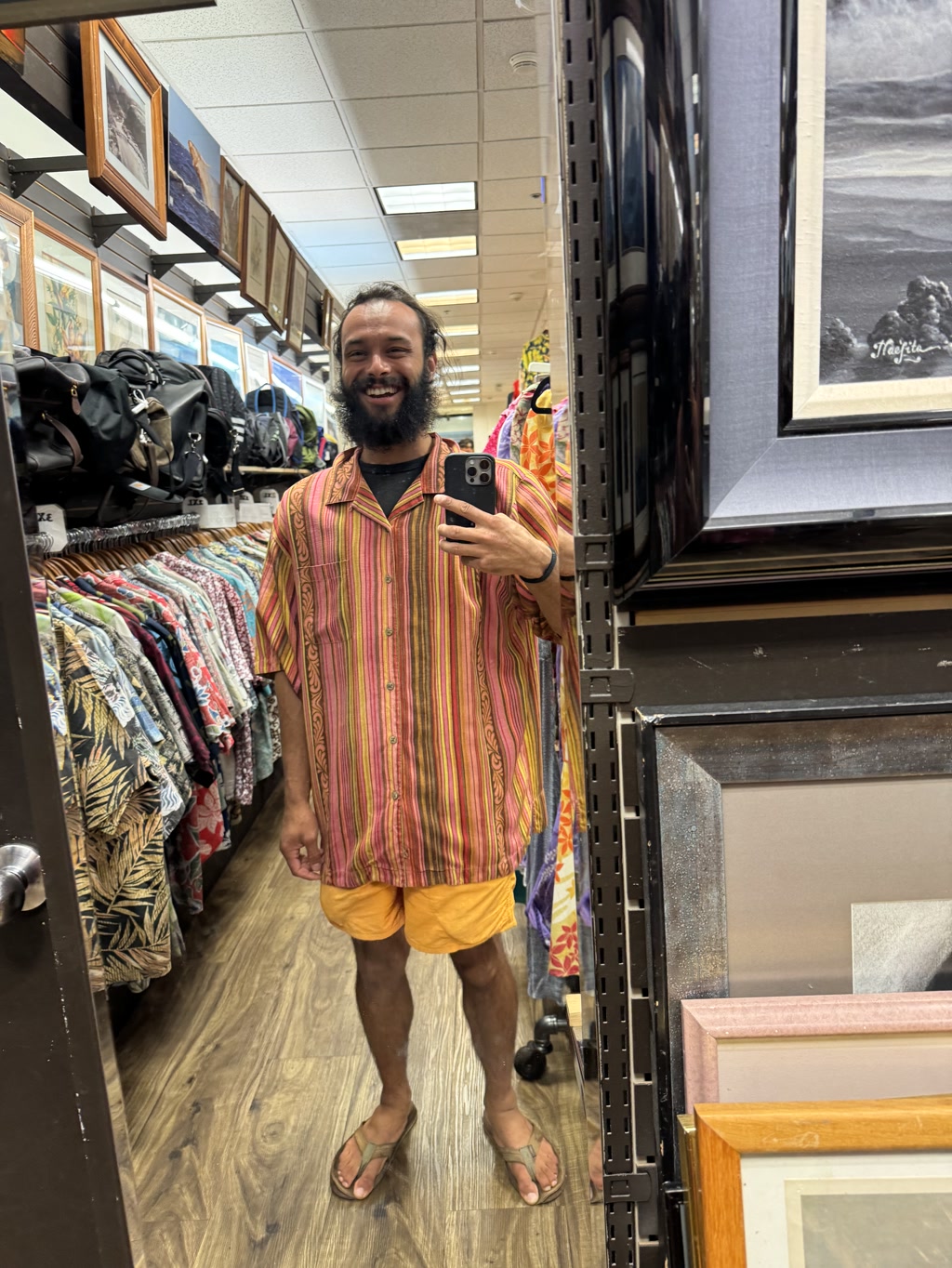 A person is standing in an aisle of a store, holding a smartphone and taking a selfie. The person is wearing a brightly colored striped shirt, yellow shorts, and sandals. The store has shelves filled with various framed pictures and racks with colorful clothing items. The setting appears to be a casual shopping environment with wooden flooring.