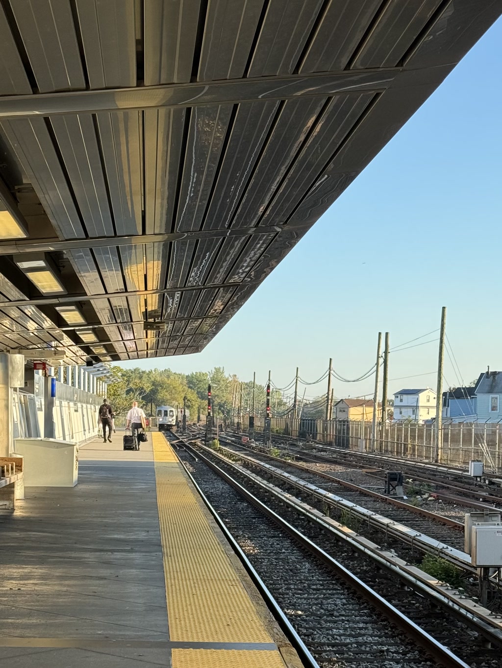 A train station platform during daytime with clear skies. The platform is equipped with benches and a tactile paving path for accessibility. Multiple rail tracks stretch into the distance, flanked by rows of poles and wires, indicative of an electrified rail system. There is some vegetation and residential buildings visible beyond the tracks on the right side. Two individuals are walking on the platform; one is pulling a suitcase. The structural features include a metal roof overhead and modern lighting fixtures. There's no visible text.