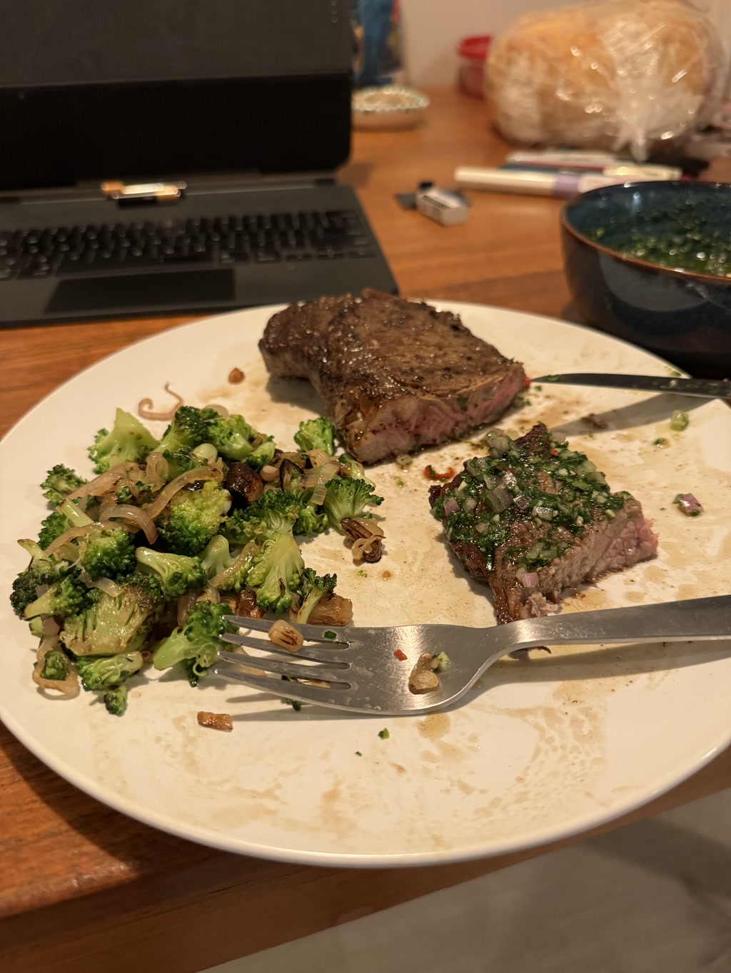 A meal consisting of a cooked steak and sautéed broccoli is on a white plate. The steak is cooked to a medium rare doneness and appears juicy with a well-seared crust sprinkled with chopped herbs. The broccoli is cooked with bits of garlic and possibly onions. The plate is on a wooden table where a laptop, a blue bowl, and various other objects such as a loaf of bread in a clear bag, pens, and a small silver object are also visible.