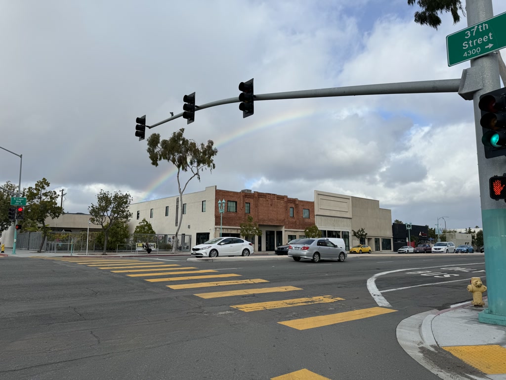 This is a view of an urban street corner. A rainbow can be seen in the sky amidst the clouds above the buildings. In the foreground, a traffic signal is illuminated with a red pedestrian stop light. The crosswalks are marked with wide yellow stripes, and various vehicles are visible on the road. There's a street sign that reads '37th street' affixed to the traffic light pole.