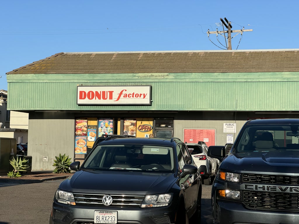 The scene includes the front view of a business establishment named 'DONUT factory' with a distinctive red signage over a green exterior. It appears to be a sunny day with clear skies. Parked in front are several vehicles, including a Volkswagen SUV in the foreground and a black Chevrolet SUV to its right. The parking lot is paved, and the establishment is single-story with a sloping roof that shows signs of weathering. There is a sign indicating 'PUBLIC PARKING' beside the entrance of the building.